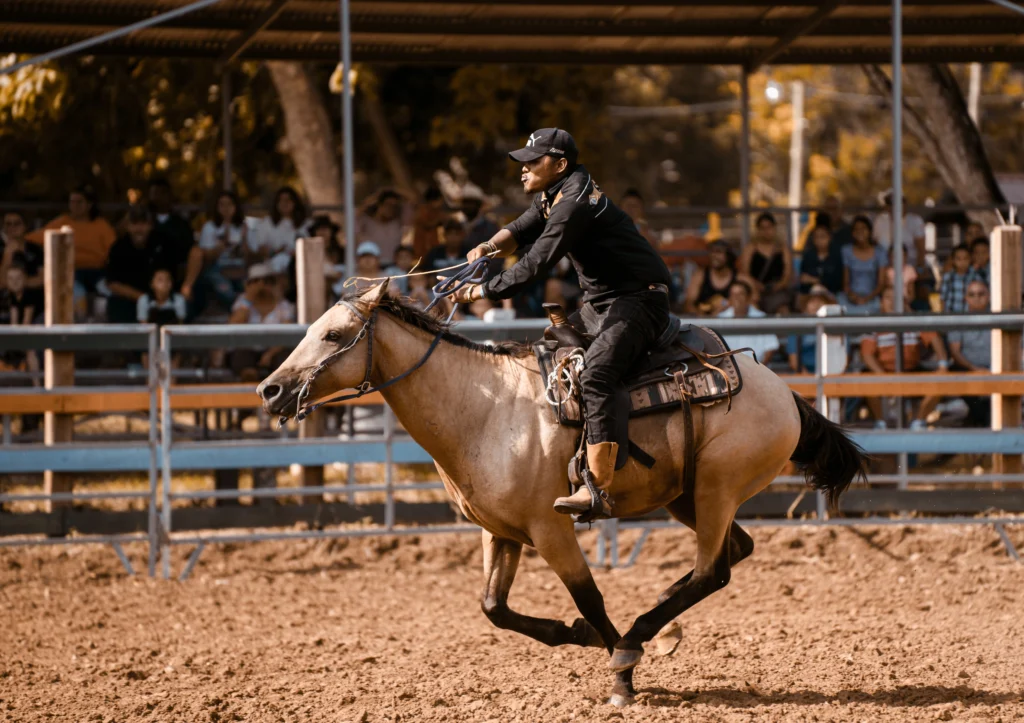 A cowboy wearing a baseball cap and black shirt rides a buckskin Quarter Horse at a fast gallop across a dirt arena. Spectators watch from the background.